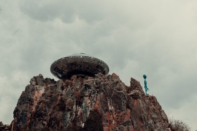 A surreal scene with a UFO and alien standing atop a rugged rock formation against a cloudy sky.