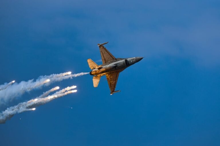 An F-16 fighter jet releases flares while performing an aerobatic maneuver against a clear blue sky at dusk.