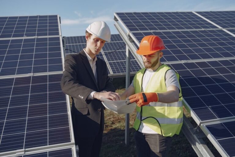 Concentrated young businessman and foreman in hardhats standing between solar panels and checking technical documentation