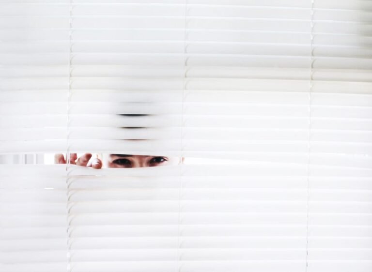 A person peeks through white blinds, creating an intriguing sense of curiosity.
