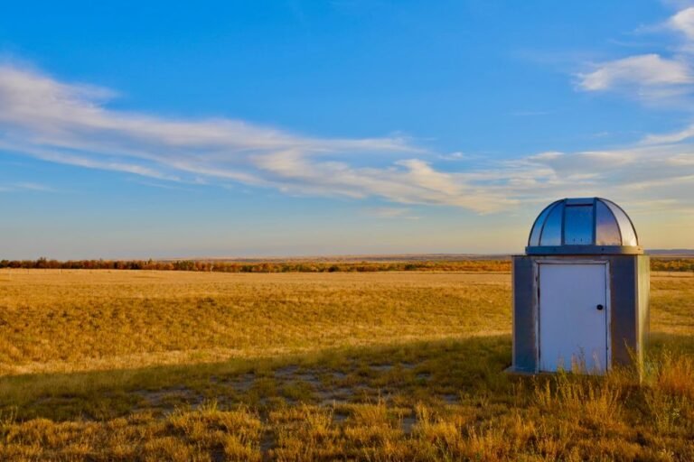 A small weather observatory building placed in a vast open grassland under a clear blue sky in Glasgow, Montana.