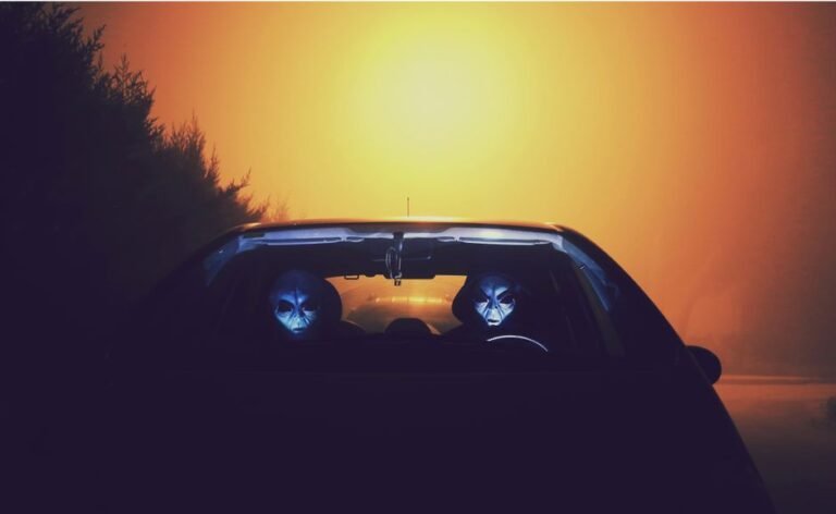 Eerie alien figures with masks seen driving a car during a colorful sunset.