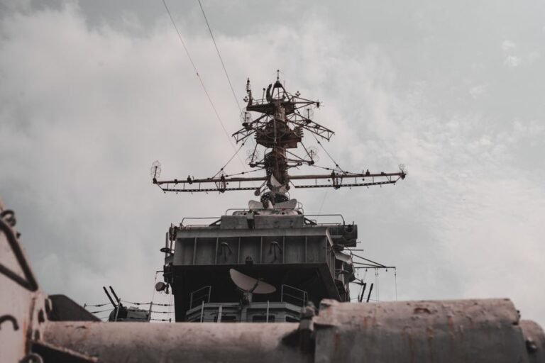 Close-up view of a military warship's radar and antenna mast against a cloudy sky.