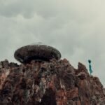 A surreal scene with a UFO and alien standing atop a rugged rock formation against a cloudy sky.