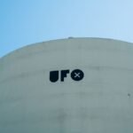 Minimalist image of a white cylindrical structure with 'UFO' text under a clear blue sky.